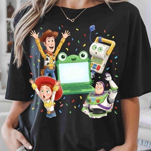 May include: A black short-sleeve t-shirt featuring a graphic print of Woody, Jessie, and Buzz Lightyear from Toy Story surrounding a green frog-shaped laptop, with colorful confetti scattered across the front of the garment.