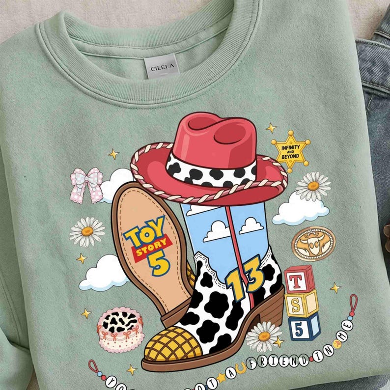 Puede incluir: Una sudadera verde salvia con un gr&aacute;fico de un sombrero de vaquero rojo y una bota con estampado de vaca. El dise&ntilde;o incluye el texto Toy Story 5, el n&uacute;mero 13 e Infinity and Beyond en una estrella, adem&aacute;s de margaritas, nubes y bloques.