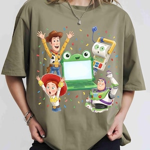 May include: An olive green t-shirt featuring a graphic print of Woody, Jessie, Buzz Lightyear, and a green frog-shaped laptop toy surrounded by colorful confetti. The characters are depicted in a celebratory pose with arms raised against the fabric.