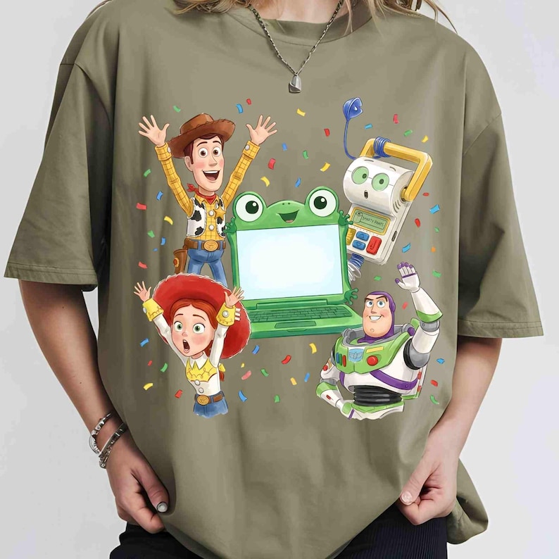 May include: An olive green t-shirt featuring a graphic print of Woody, Jessie, Buzz Lightyear, and a green frog-shaped laptop toy surrounded by colorful confetti. The characters are depicted in a celebratory pose with arms raised against the fabric.