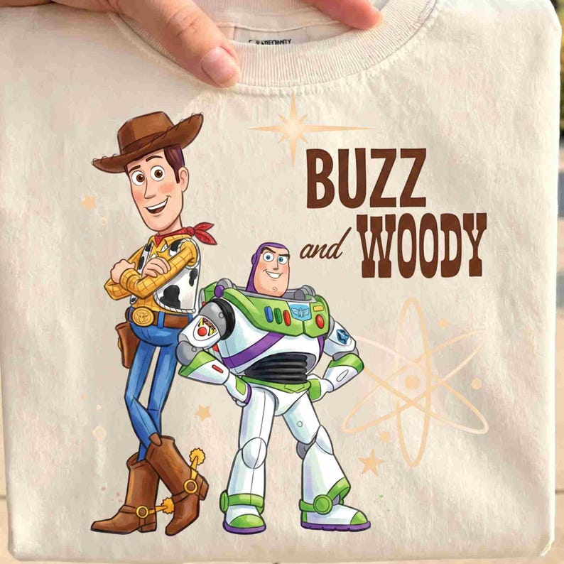 May include: A cream-colored t-shirt featuring a graphic print of the characters Woody and Buzz Lightyear. Woody wears a brown cowboy hat, yellow shirt, and blue jeans. Buzz wears a white, green, and purple space suit. The text reads BUZZ and WOODY.