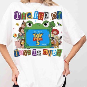 Pode incluir: Uma t-shirt branca com uma impress&atilde;o gr&aacute;fica de v&aacute;rias personagens de Toy Story rodeando uma moldura verde em forma de sapo. O texto diz THE AGE OF TOY STORY 5 ONLY IN THEATERS JUNE 19 TOYS IS OVER em letras coloridas estilo nota de resgate.