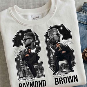 May include: A white crewneck sweatshirt featuring a large graphic print of the number 26. Inside the numbers are black and white portraits of Usher Raymond and Chris Brown, city skylines, and star accents. The text reads Usher Raymond and Chris Brown.