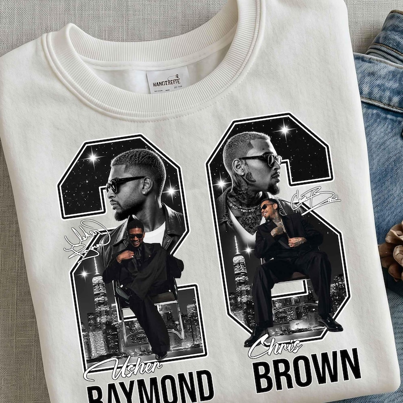 May include: A white crewneck sweatshirt featuring a large graphic print of the number 26. Inside the numbers are black and white portraits of Usher Raymond and Chris Brown, city skylines, and star accents. The text reads Usher Raymond and Chris Brown.
