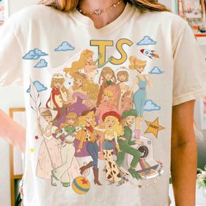 May include: A cream-colored t-shirt featuring a graphic print of multiple animated characters in various outfits, including a space suit, cowgirl attire, and gowns. The design includes the letters TS, a rocket, clouds, and a star-shaped sheriff badge.