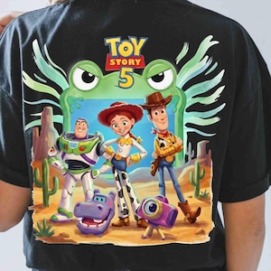 May include: A black t-shirt featuring a Toy Story 5 graphic. The design displays Buzz Lightyear, Jessie, and Woody standing in a desert landscape with a purple hippo and a camera. A large green frog face is positioned behind the characters.