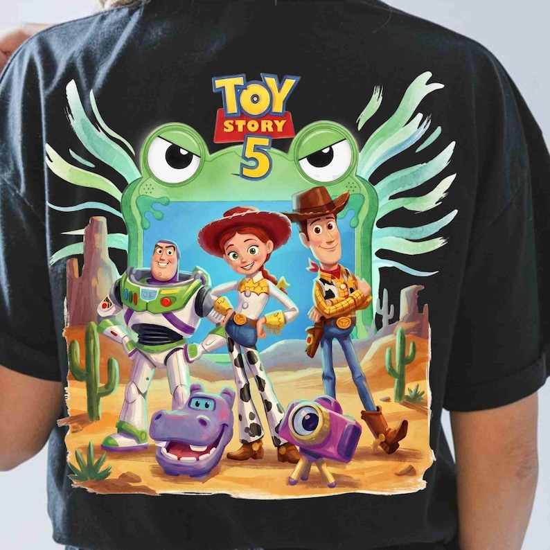 May include: A black t-shirt featuring a Toy Story 5 graphic. The design displays Buzz Lightyear, Jessie, and Woody standing in a desert landscape with a purple hippo and a camera. A large green frog face is positioned behind the characters.