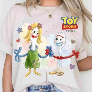 May include: A light-colored t-shirt featuring a graphic print of the Toy Story characters Forky and Karen Beverly holding hands. The design includes watercolor-style splashes, small stars, and hearts, with the Toy Story Pixar logo printed in the upper right.