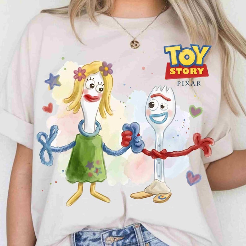 May include: A light-colored t-shirt featuring a graphic print of the Toy Story characters Forky and Karen Beverly holding hands. The design includes watercolor-style splashes, small stars, and hearts, with the Toy Story Pixar logo printed in the upper right.