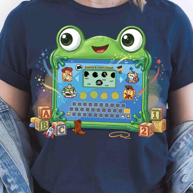 May include: A dark blue t-shirt featuring a graphic of a green frog-shaped tablet with the text Explore & Learn Lilypad. The screen displays characters from Toy Story, a keyboard, and various icons. Toy blocks and a cowboy boot are below.