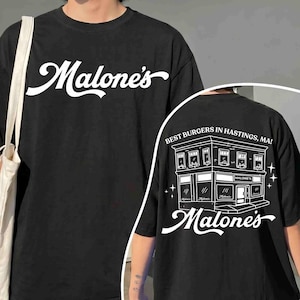 May include: Black t-shirt with the white text "Malone's" on the front. The back of the shirt features a white illustration of a building with the text "Best Burgers in Hastings, MA!" and "Malone's".