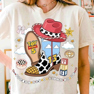 Puede incluir: Una camiseta color crema con un gr&aacute;fico de un sombrero de vaquero y una bota con estampado de vaca. El dise&ntilde;o incluye el texto Toy Story 5, el n&uacute;mero 13 y You've Got A Friend In Me, junto a ilustraciones de margaritas, nubes y bloques.