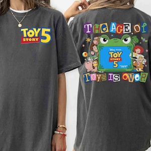 Pode incluir: Uma t-shirt cinzento-carv&atilde;o com um pequeno log&oacute;tipo Toy Story 5 no peito esquerdo. As costas apresentam um gr&aacute;fico grande com uma moldura de sapo verde, v&aacute;rias personagens de Toy Story e o texto The Age of Toys is Over em letras coloridas.