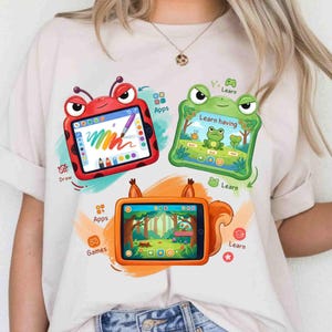 May include: A cream-colored t-shirt featuring a graphic print of three cartoon tablets. One is a red ladybug, one is a green frog, and one is an orange squirrel. Each tablet displays colorful digital interfaces with text including Apps, Draw, Learn, and Games.