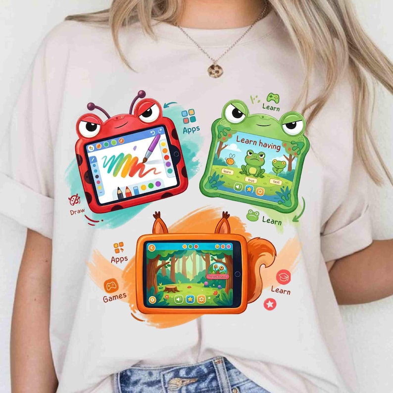 May include: A cream-colored t-shirt featuring a graphic print of three cartoon tablets. One is a red ladybug, one is a green frog, and one is an orange squirrel. Each tablet displays colorful digital interfaces with text including Apps, Draw, Learn, and Games.