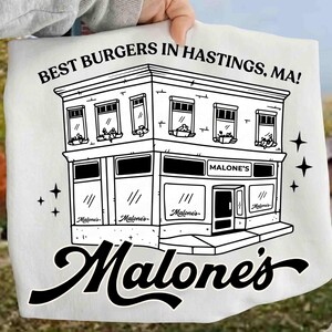 May include: White t-shirt with a black illustration of a two-story building with the text "BEST BURGERS IN HASTINGS, MA!" and "Malone's" in a cursive font. The building has multiple windows and a door.