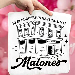 May include: A white tote bag featuring a black and white illustration of a two-story building with the text "BEST BURGERS IN HASTINGS, MA!" and "Malone's" in a stylized font. The design includes stars.