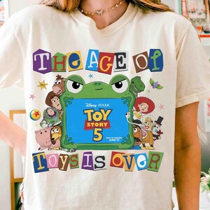 May include: A cream-colored t-shirt featuring a graphic of a green frog frame surrounding the text Disney Pixar Toy Story 5 In Theaters June 19. The design includes various animated characters and the text The Age Of Toys Is Over in colorful letters.