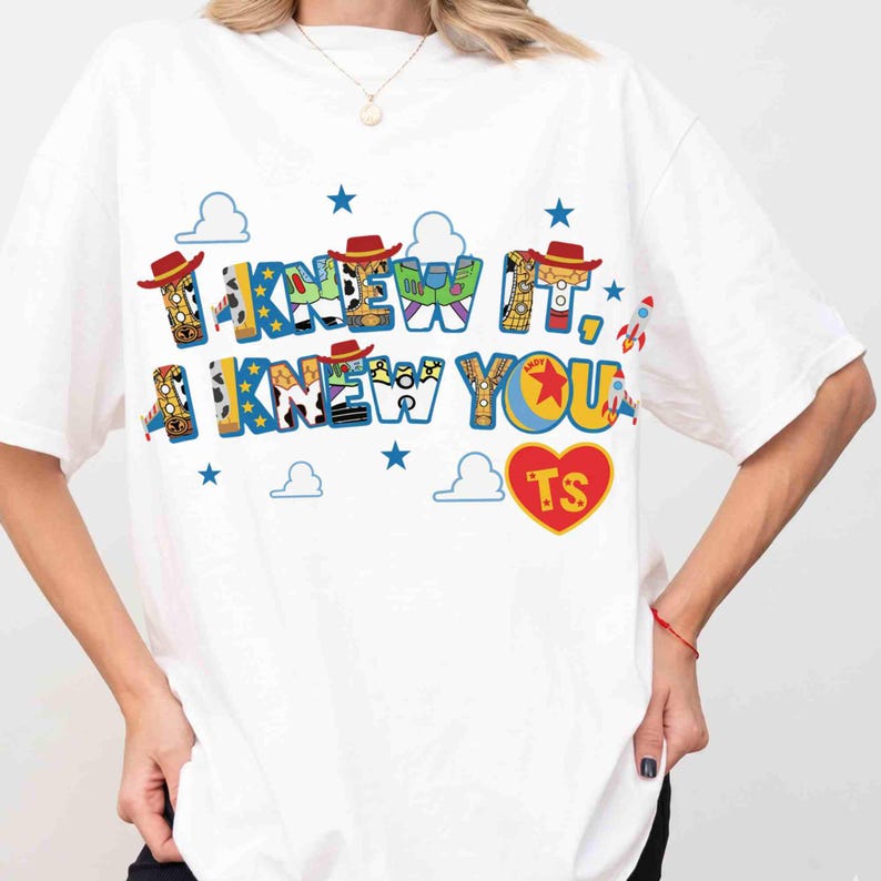 May include: A white short-sleeved t-shirt featuring a graphic print with the text I KNEW IT, I KNEW YOU in blue letters filled with patterns inspired by toy characters. The design includes small red cowboy hats, blue stars, white clouds, rockets, and a red heart with the letters TS.