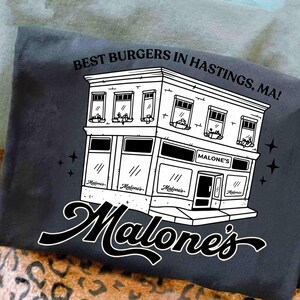 May include: Dark gray t-shirt featuring a black and white illustration of a building with the text "BEST BURGERS IN HASTINGS, MA!". The building has the name "Malone's" on it. The shirt has a casual style.