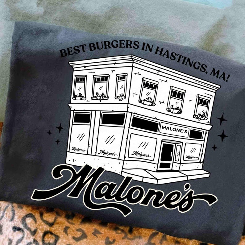 May include: Dark gray t-shirt featuring a black and white illustration of a building with the text "BEST BURGERS IN HASTINGS, MA!". The building has the name "Malone's" on it. The shirt has a casual style.