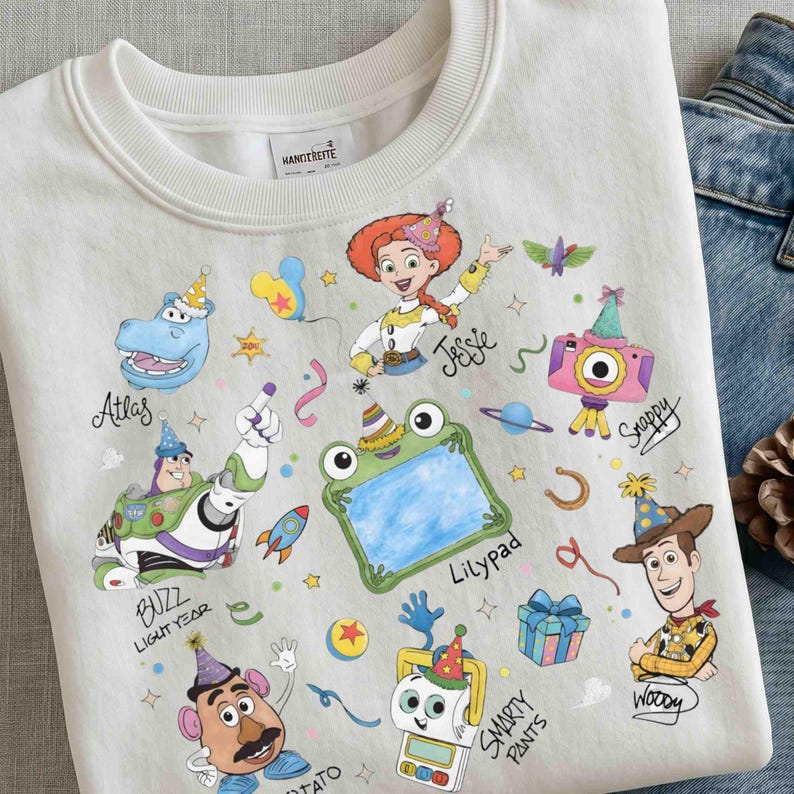 May include: A white crewneck sweatshirt featuring a colorful graphic print of various cartoon characters wearing party hats. The design includes Buzz Lightyear, Jessie, Woody, Mr. Potato Head, and a frog, with handwritten names and festive icons.