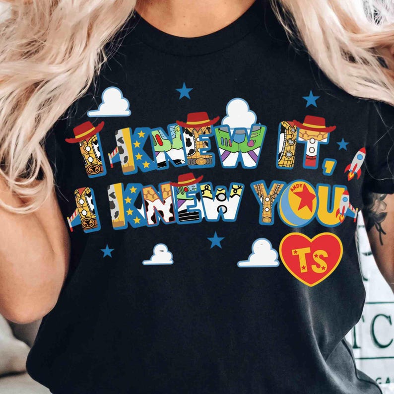 May include: A black t-shirt featuring the text I KNEW IT, I KNEW YOU in blue block letters filled with patterns inspired by Toy Story characters. The design includes small red cowboy hats, white clouds, blue stars, rockets, and a red heart with TS.