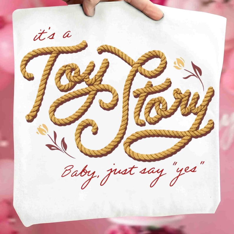 Puede incluir: Una superficie de tela blanca con el texto "it's a Toy Story" escrito en una fuente de textura de cuerda color canela. Debajo, el texto "Baby, just say "yes"" est&aacute; escrito en cursiva roja. Incluye dos peque&ntilde;as ilustraciones florales en rojo y canela.