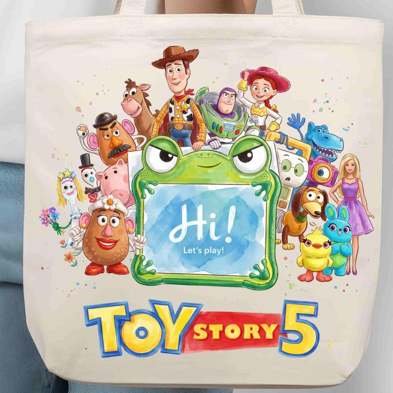May include: A cream-colored canvas tote bag featuring a colorful graphic of various Toy Story characters surrounding a green frog-shaped frame with the text Hi! Let's play! and the large blue and red text Toy Story 5 printed at the bottom.
