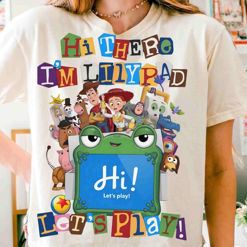 Puede incluir: Una camiseta de color crema con un estampado gr&aacute;fico de varios personajes de Toy Story. El texto "Hi There I'm Lilypad" aparece en la parte superior y "Let's Play!" en la inferior. Una tableta verde con forma de rana muestra "Hi! Let's play!" en el centro.