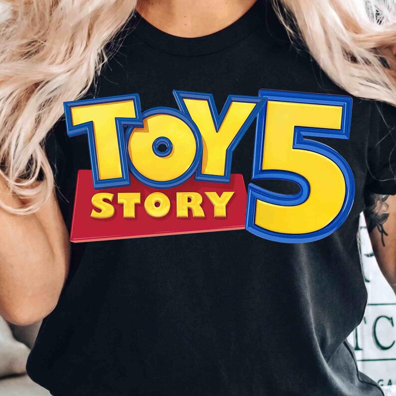 Pode incluir: Uma t-shirt preta de manga curta com uma grande impress&atilde;o gr&aacute;fica na frente. O design apresenta o texto TOY STORY em letras amarelas em negrito com contornos azuis, posicionado acima de um grande n&uacute;mero 5 amarelo sobre um fundo vermelho.