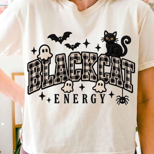 May include: A cream-colored short-sleeve t-shirt featuring a graphic print with the text BLACK CAT ENERGY in a black and white buffalo plaid pattern. The design includes a black cat, white ghosts, black bats, a spider, and small stars.