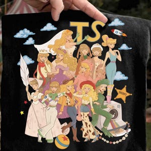 May include: A black t-shirt featuring a graphic print of multiple blonde characters in various outfits, including a space suit, cowgirl attire, and formal gowns. The design includes blue clouds, a rocket, a yellow star badge labeled SHERIFY, and the letters TS.