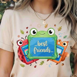May include: A beige t-shirt featuring a graphic of a green frog, a red ladybug, and an orange squirrel, each holding a blue tablet. The central frog tablet displays the text "best friends" in cursive script, surrounded by small leaves and acorns.