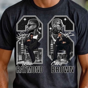 May include: A charcoal gray t-shirt featuring a graphic print of Usher Raymond and Chris Brown. The design includes two large numbers, portraits of the artists wearing sunglasses, city skylines, star patterns, and their names printed in white.