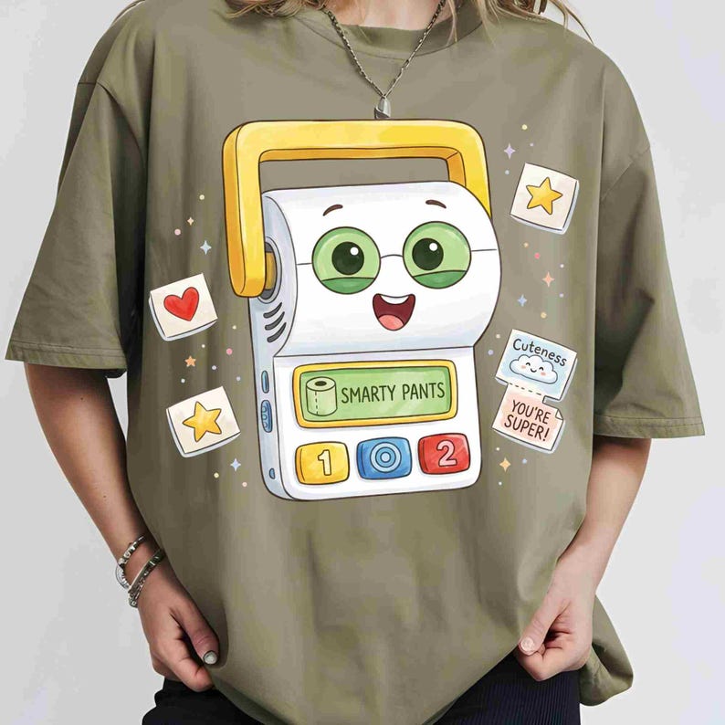 May include: An olive green t-shirt featuring a cartoon graphic of a white label maker with a yellow handle, green eyes, and a screen displaying the text "SMARTY PANTS". The design includes small icons of hearts, stars, and text bubbles.