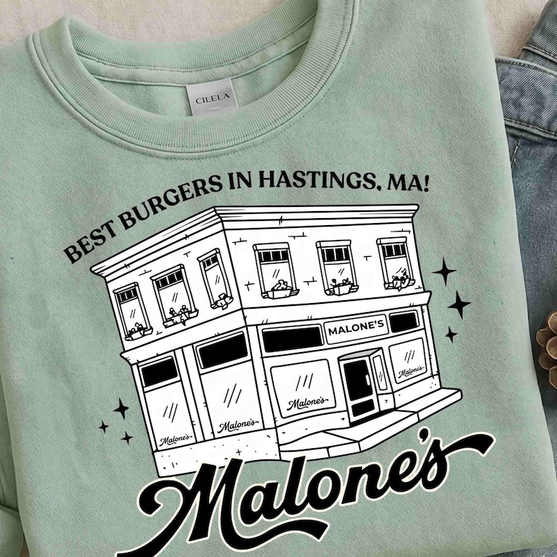 May include: A light green sweatshirt featuring a black and white illustration of a building with the text "BEST BURGERS IN HASTINGS, MA!" and "Malone's". The design includes stars and the brand name.