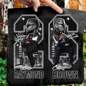May include: A black fabric item featuring a graphic print of Usher Raymond and Chris Brown. Each artist is depicted in a large number 23 frame with a city skyline background, wearing sunglasses and a suit. The names are printed in white text below.