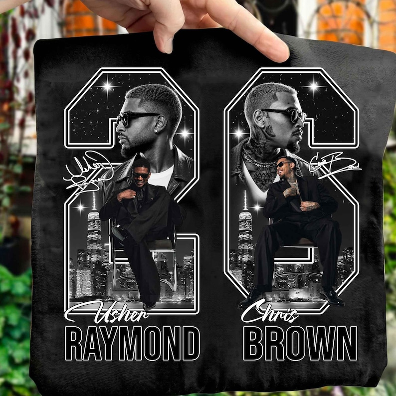 May include: A black fabric item featuring a graphic print of Usher Raymond and Chris Brown. Each artist is depicted in a large number 23 frame with a city skyline background, wearing sunglasses and a suit. The names are printed in white text below.