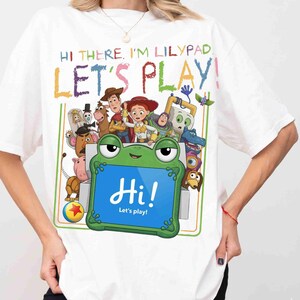 May include: A white t-shirt featuring a colorful graphic of Toy Story characters and a green frog-shaped tablet. The text reads: HI THERE. I'M LILYPAD. LET'S PLAY! and Hi! Let's play! The design includes Woody, Jessie, Buzz Lightyear, and others.