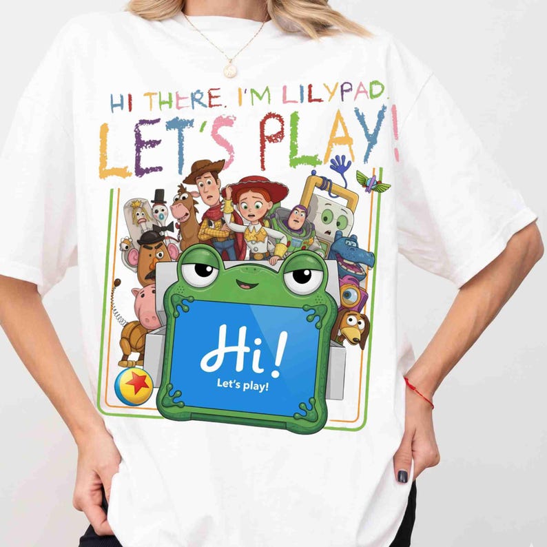 May include: A white t-shirt featuring a colorful graphic of Toy Story characters and a green frog-shaped tablet. The text reads: HI THERE. I'M LILYPAD. LET'S PLAY! and Hi! Let's play! The design includes Woody, Jessie, Buzz Lightyear, and others.