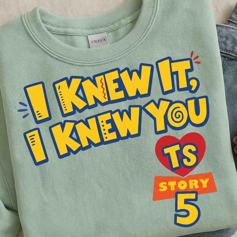 May include: A light green crewneck sweatshirt featuring the yellow text I KNEW IT, I KNEW YOU with blue outlines. Below the text is a red heart containing the letters TS, followed by the word STORY and the number 5 in yellow with blue outlines.
