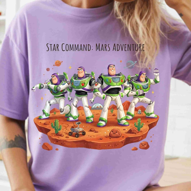 May include: A light purple t-shirt featuring a graphic of four Buzz Lightyear figures on a red Martian landscape. The text "STAR COMMAND: MARS ADVENTURE" is printed above the characters, who are surrounded by small cacti, rocks, and a rover.