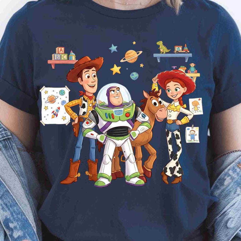 May include: A navy blue short-sleeved t-shirt featuring a colorful graphic print of Woody, Buzz Lightyear, Bullseye, and Jessie from Toy Story. The design includes floating planets, stars, toy blocks, and children's drawings on a dark blue background.