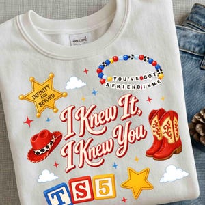 May include: A white crewneck sweatshirt featuring a graphic print with the text I KNEW IT, I KNEW YOU, a yellow sheriff star badge labeled INFINITY AND BEYOND, a red cowboy hat, a red cowboy boot, a beaded bracelet, and colorful alphabet blocks.