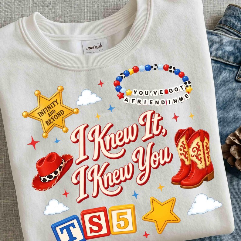 May include: A white crewneck sweatshirt featuring a graphic print with the text I KNEW IT, I KNEW YOU, a yellow sheriff star badge labeled INFINITY AND BEYOND, a red cowboy hat, a red cowboy boot, a beaded bracelet, and colorful alphabet blocks.
