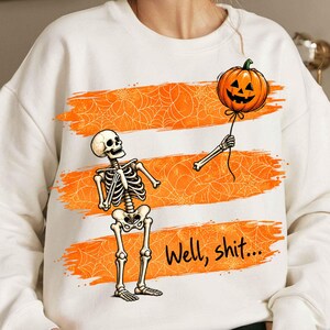 Könnte beinhalten: Ein cremefarbenes Sweatshirt mit einer Grafik eines Skeletts, das einen schwebenden Kürbis-Ballon betrachtet, der von einem abgetrennten Skelettarm gehalten wird. Das Design zeigt orangefarbene Pinselstriche mit Spinnennetzmustern und den Text Well, shit...