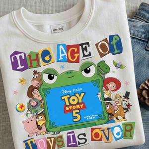Pode incluir: Uma camisola branca de gola redonda com uma impress&atilde;o gr&aacute;fica colorida com o texto THE AGE OF TOY STORY 5 TOYS IS OVER. O design inclui um sapo verde grande a segurar um sinal azul e v&aacute;rias personagens animadas do filme.