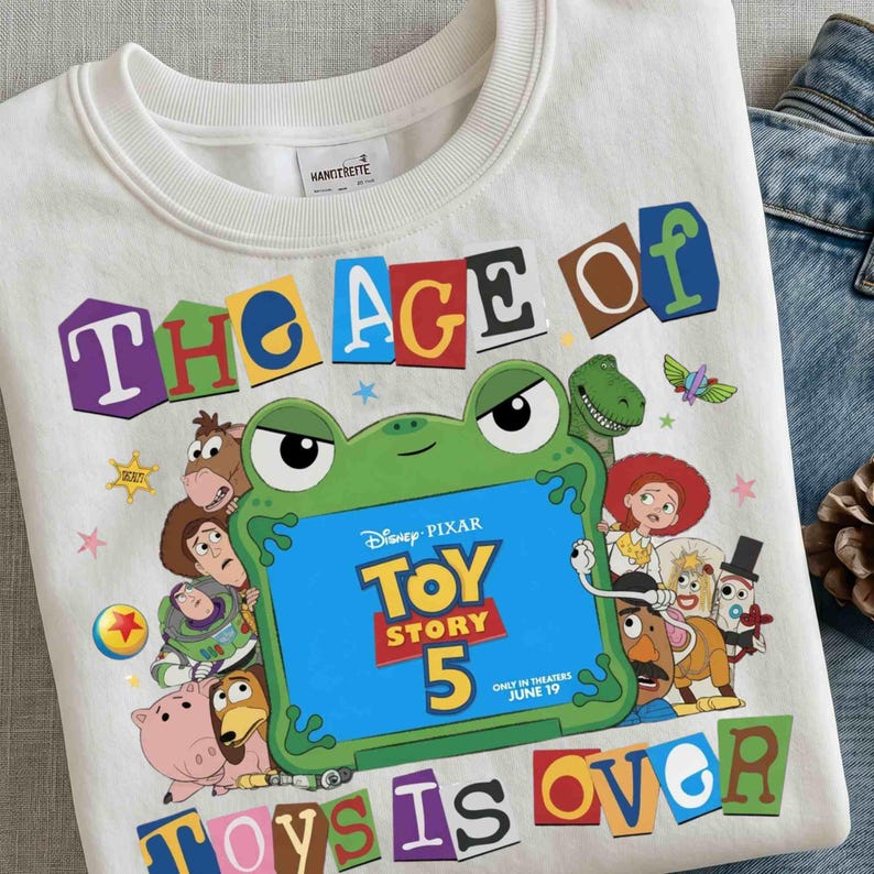 Pode incluir: Uma camisola branca de gola redonda com uma impress&atilde;o gr&aacute;fica colorida com o texto THE AGE OF TOY STORY 5 TOYS IS OVER. O design inclui um sapo verde grande a segurar um sinal azul e v&aacute;rias personagens animadas do filme.