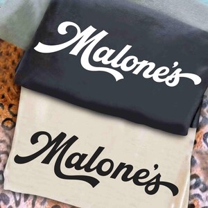May include: Two folded t-shirts, one black and one beige, are stacked. Each shirt features the word "Malone's" in a cursive font. The black shirt has white text, while the beige shirt has black text. The shirts are on a patterned surface.